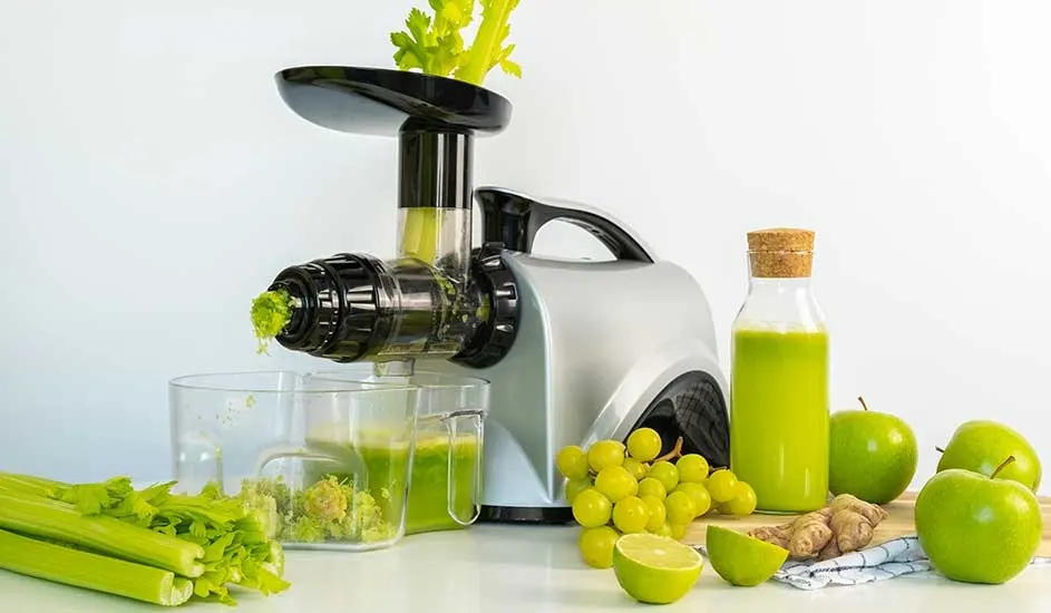 A silver juicer presses fresh celery juice beside green apples, grapes, lime and ginger on a neat white kitchen counter