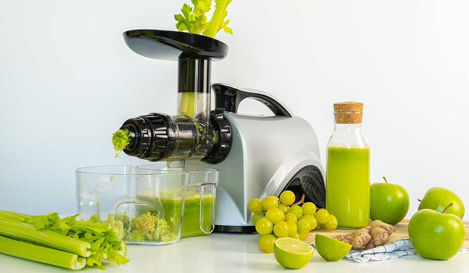 Op de afbeelding staat een zilveren slowjuicer van Omega, omringd door bleekselderij, groene appels, druiven, limoen en een fles groentesap.