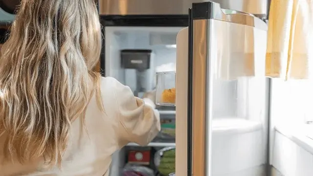 A woman with long blonde hair is reaching into a silver Samsung fridge in a bright kitchen, searching for something on the middle shelf.