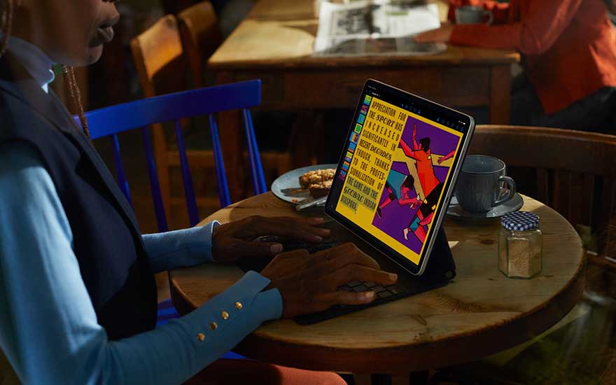 Persoon werkt in een café aan een tablet met toetsenbordhoes op ronde houten tafel, kleurrijke illustratie op het scherm naast koffie en snack.