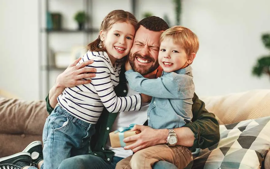 Een blije vader wordt op de bank stevig geknuffeld door twee lachende kinderen, terwijl hij een klein cadeau vasthoudt.