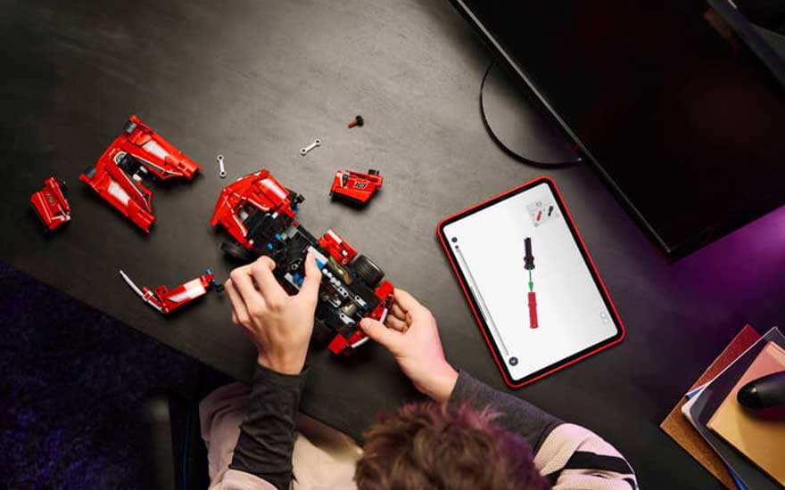 Bovenaanzicht van handen die een rode LEGO-sportwagen bouwen, naast losse onderdelen en een tablet met instructies.