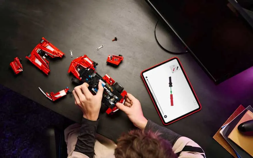 Bovenaanzicht van handen die een rode LEGO-sportwagen bouwen, naast losse onderdelen en een tablet met instructies.