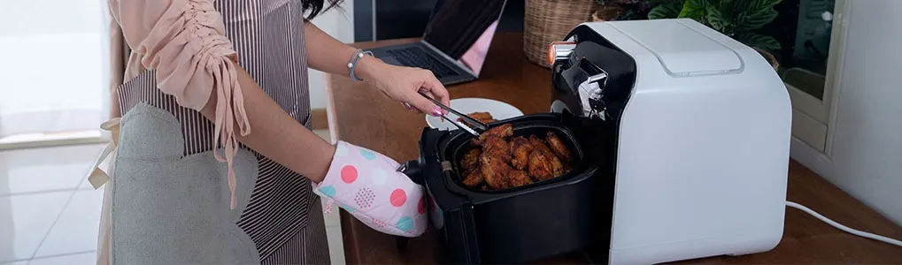Person in an apron uses tongs to remove food from a black air fryer next to a laptop on a wooden table.