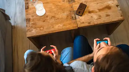 Vanuit vogelperspectief: twee mensen spelen games. Op een houten tafel staat een glas water en een telefoon.