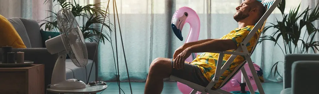Man relaxing in a beach chair indoors with a pink flamingo floatie behind him. A fan and plants are also visible.