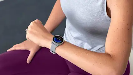 Een vrouw met een grijze sporttop en paarse legging draagt een grijze Garmin smartwatch, zittend op een fitnessbal of bank.