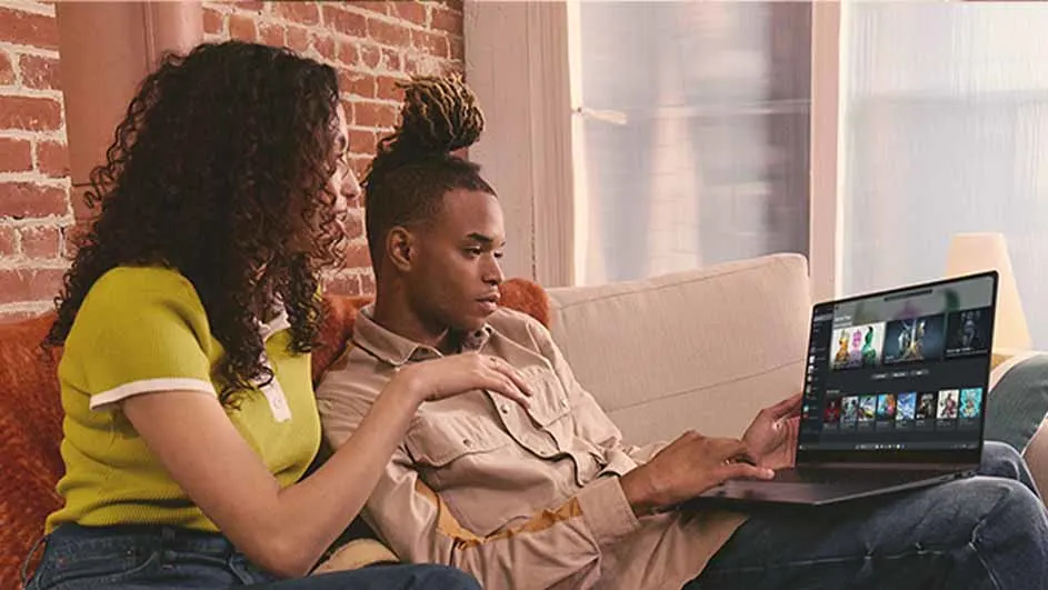 Een man en vrouw zitten op een bank en kijken naar een laptop met een streamingdienst. Achter hen is een bakstenen muur.