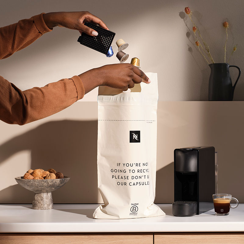 Op een aanrecht staat een zak met koffiecapsules, een koffiezetapparaat, een kopje espresso en een schaal met walnoten.