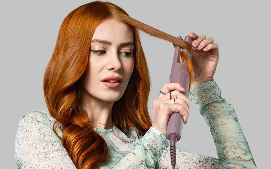 Young woman with long copper‑red hair uses a purple curling styler, wrapping a thin section around the heated barrel while looking thoughtfully aside.