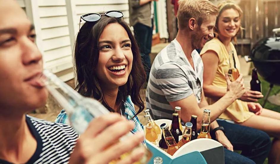 Friends laugh and drink together outdoors at a barbecue; bottles sit in a cool box while the mood feels warm and relaxed.
