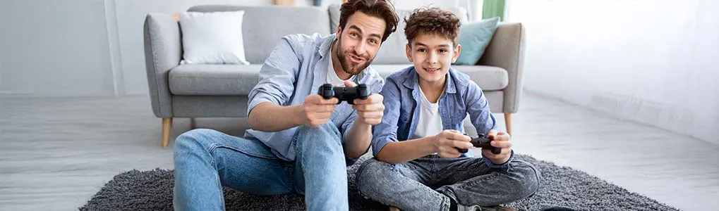 Twee personen, een volwassene en een kind, zitten op de vloer in een woonkamer en spelen samen een videogame met controllers in de hand.