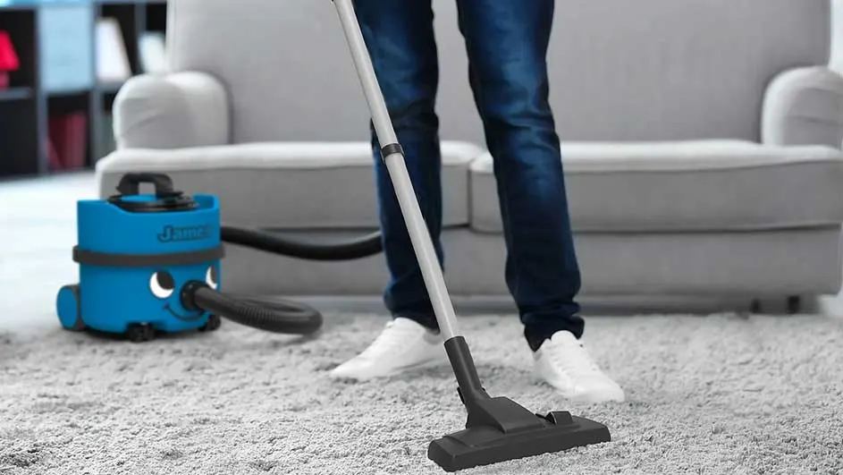 Een persoon stofzuigt een grijze vloerkleed met een blauwe stofzuiger. Een grijze bank staat op de achtergrond.