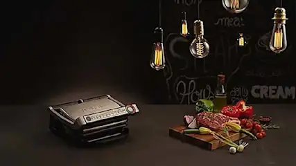 Een elektrische grill links, een houten plank met groenten en vlees rechts, en hangende lampen tegen een donkere achtergrond.