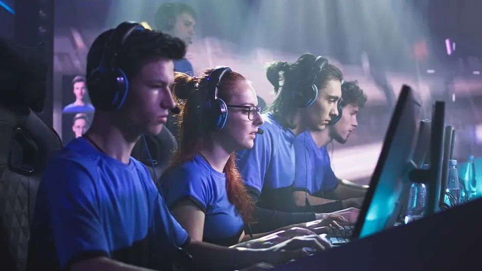 Esports team in blue jerseys sits focused at PCs with headsets, working together in an intense match on a brightly lit stage.