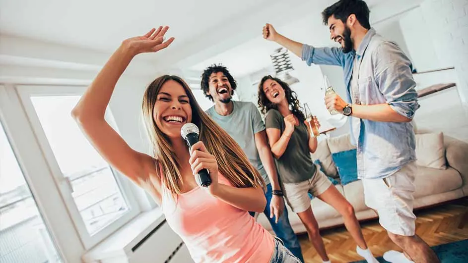 Een groep van vier vrienden zingt karaoke in een woonkamer, met een microfoon in de hand en drankjes in de andere.