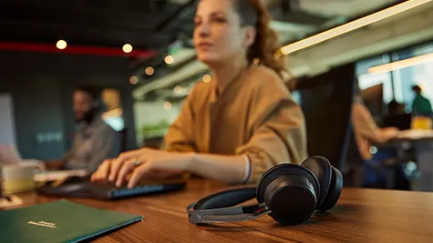 Hoofdtelefoon ligt op houten bureau in open kantoor, terwijl een collega op de achtergrond achter haar computer werkt.