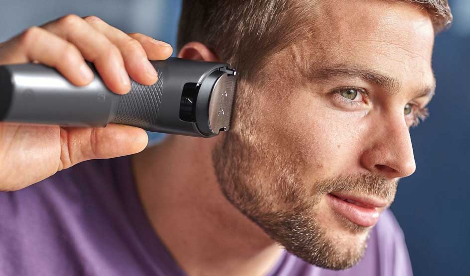 A man trims his hair near the ear with an electric device; his face is clearly visible in soft light.