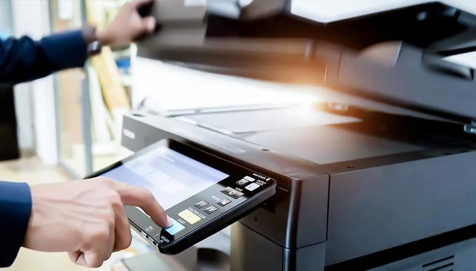 Een persoon in een blauw jasje bedient een zwarte printer met een digitaal scherm in een kantooromgeving.