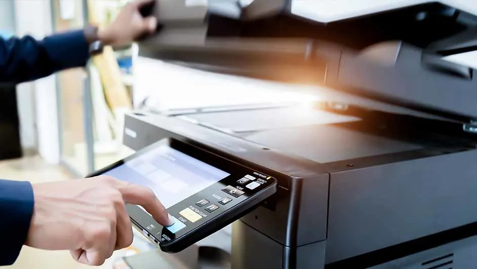 Een persoon in een blauw jasje bedient een zwarte printer met een digitaal scherm in een kantooromgeving.