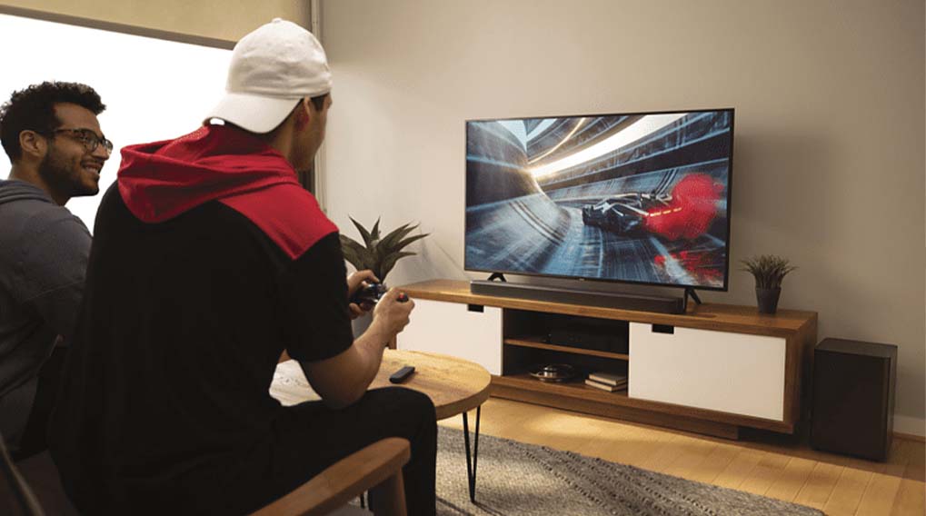 Twee mannen kijken naar een racegame op een tv, geplaatst op een houten kast met witte deuren en planten.