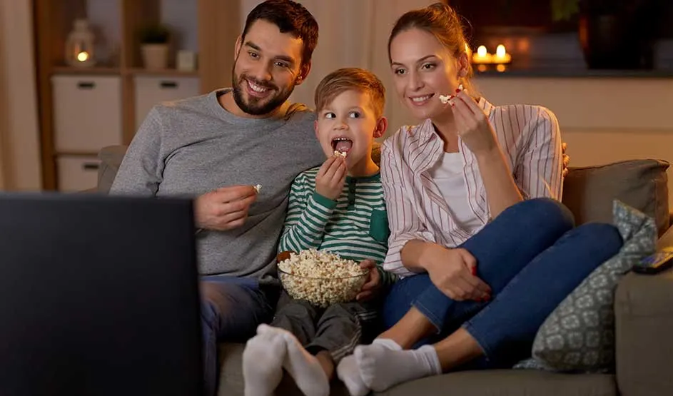 Een gezin zit gezellig op de bank met een grote kom popcorn en kijkt samen televisie in een sfeervolle woonkamer.