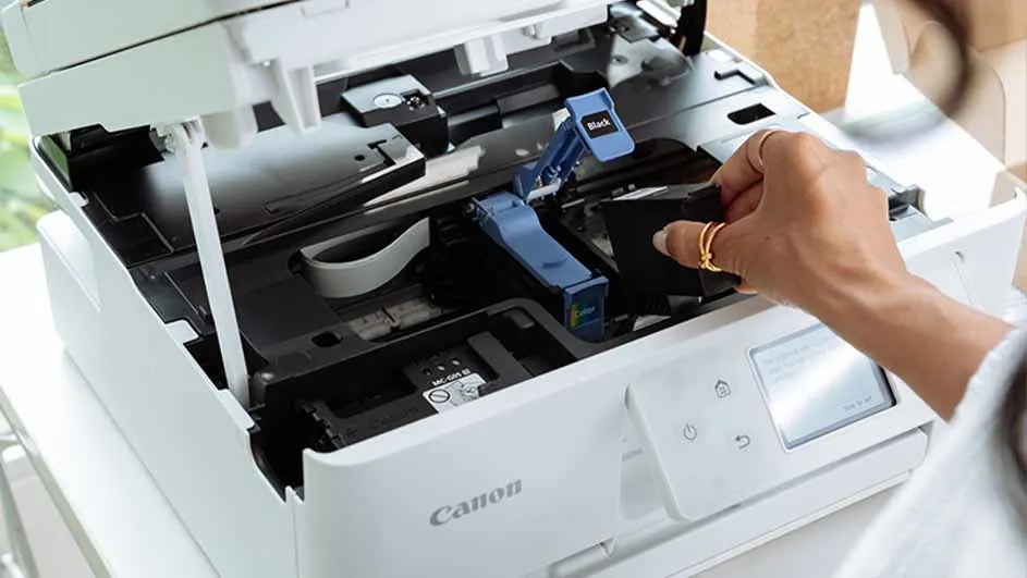 Een hand met ringen plaatst een zwarte inktcartridge in een openstaande, witte Canon printer. Blauwe clips zijn zichtbaar.