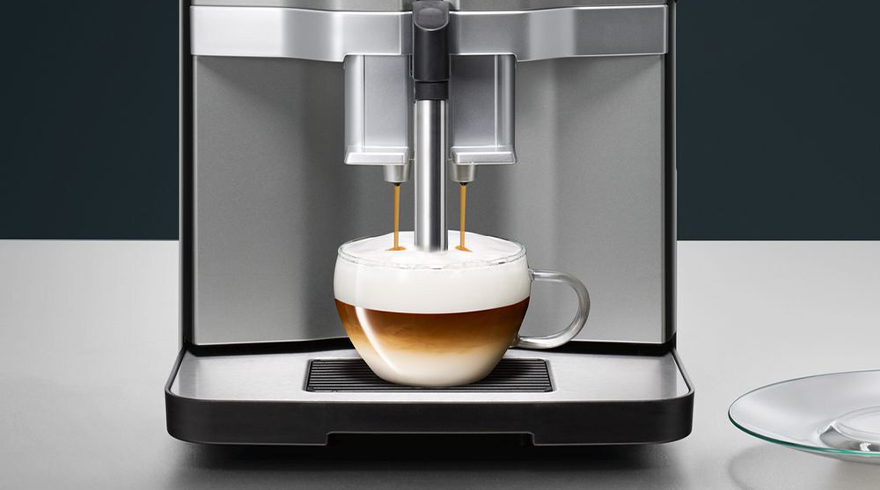 Een koffiemachine vult een glazen kop met koffie en melkschuim, op een witte tafel tegen een donkere achtergrond.