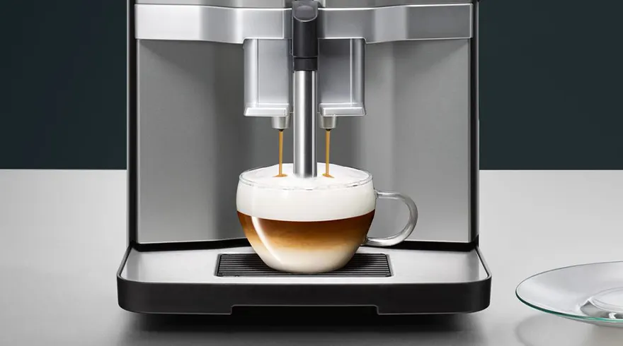 Een koffiemachine vult een glazen kop met koffie en melkschuim, op een witte tafel tegen een donkere achtergrond.
