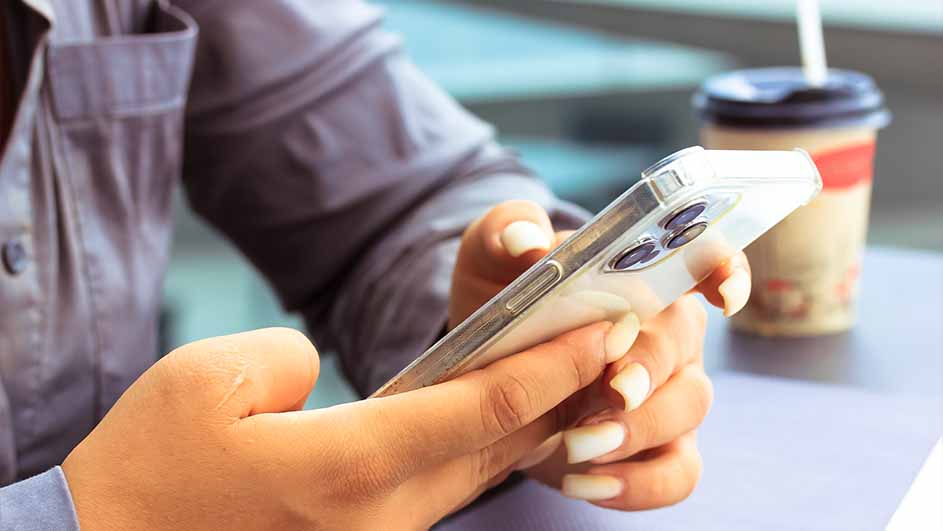 Een persoon houdt een zilverkleurige smartphone vast, op de achtergrond een beker met rietje.