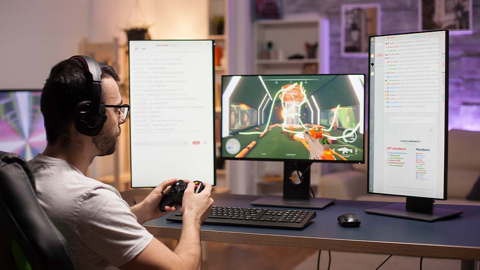 Man speelt computerspel met controller op desktop met drie monitoren, toetsenbord en muis.