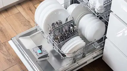 Een open vaatwasser gevuld met witte borden, kommen en zilverwerk in een metalen rek. De vaatwasser heeft een zilveren binnenkant.