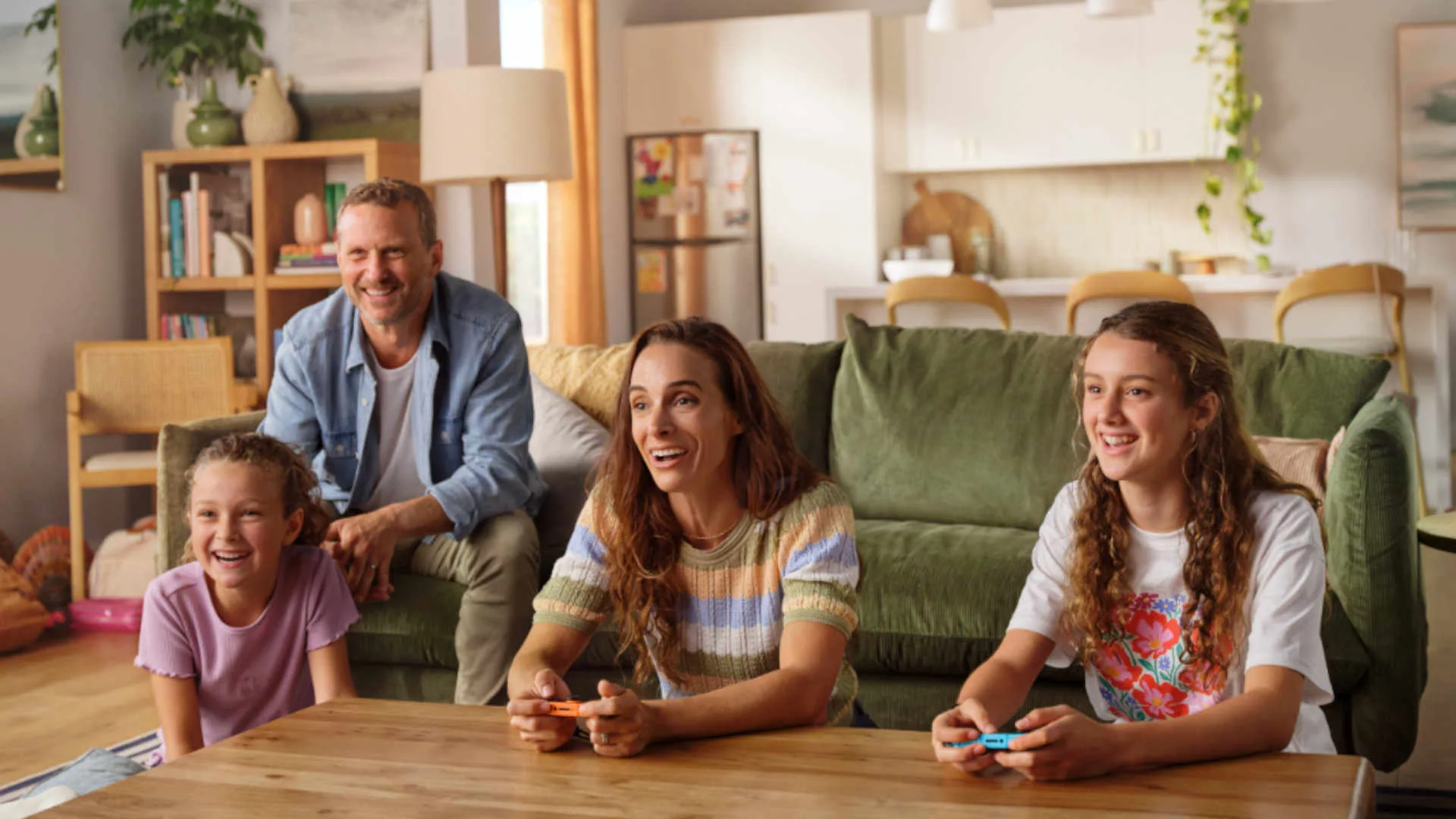 Vier mensen, twee volwassenen en twee kinderen, zitten lachend op een bank en spelen samen een videospelletje in een gezellige woonkamer.