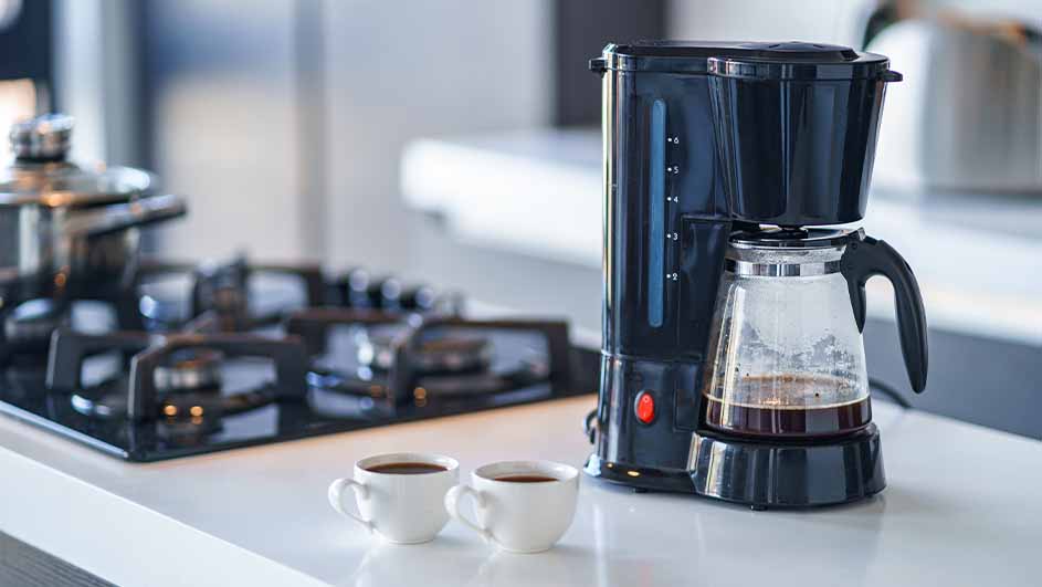 Black filter coffee machine with a glass jug containing brewed coffee, placed on a white kitchen counter next to two small cups of coffee and a gas stove.