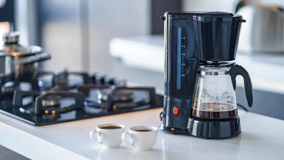 Black filter coffee machine with a glass jug containing brewed coffee, placed on a white kitchen counter next to two small cups of coffee and a gas stove.