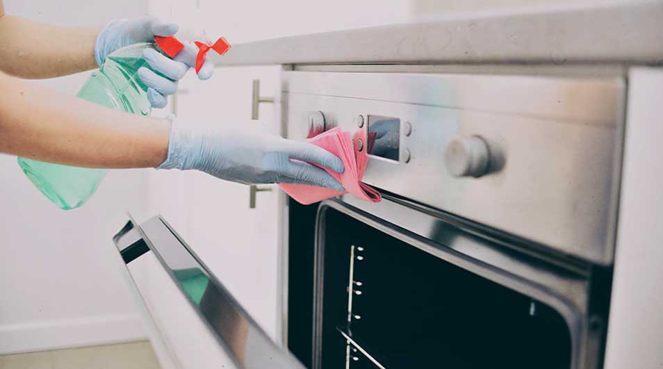 Someone wearing gloves cleans a stainless-steel oven with a pink cloth and a spray bottle.