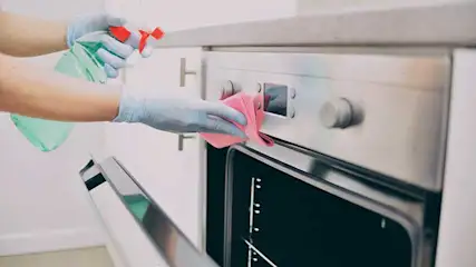 Iemand met handschoenen maakt een roestvrijstalen oven schoon met een roze doek en een spuitfles.
