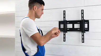 Een man in een blauwe overall monteert een zwarte tv-beugel op een witte houten wand.