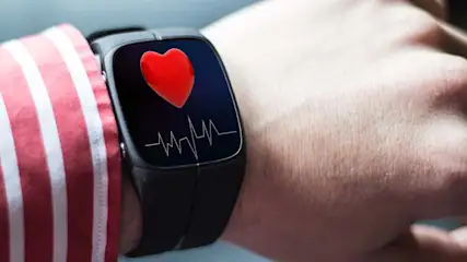 Een polshorloge met een hartslagmeter en een rode hartpictogram op een pols naast een rood-wit gestreept overhemd.