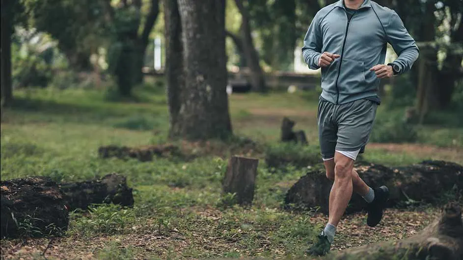 Een persoon in grijze sportkleding rent door een bosrijk gebied met groene bomen en grond.