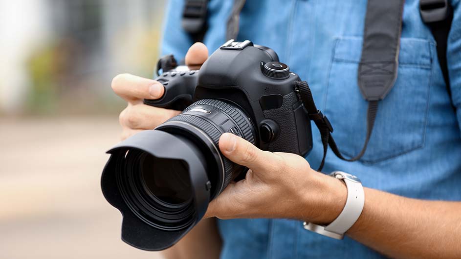 Een persoon houdt een zwarte camera met een lens vast, en draagt een blauw overhemd en een horloge met een witte band.