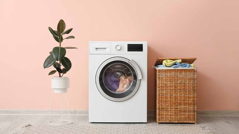 A laundry room setting with a white washing machine, wicker basket with clothes, and a potted plant against a peach wall.