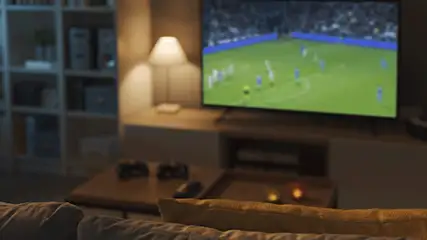 Woonkamer met een tv waarop een voetbalwedstrijd te zien is, een lamp, een bank en een kast.