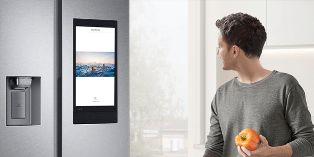 Man met oranje paprika in keuken, naast koelkast met scherm waarop een foto van een zee staat.