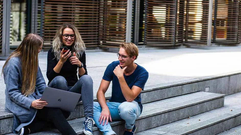 Drie mensen zitten op een trap. Eén gebruikt een laptop, één een telefoon en de ander kijkt toe. Achtergrond: gebouw met horizontale lijnen.