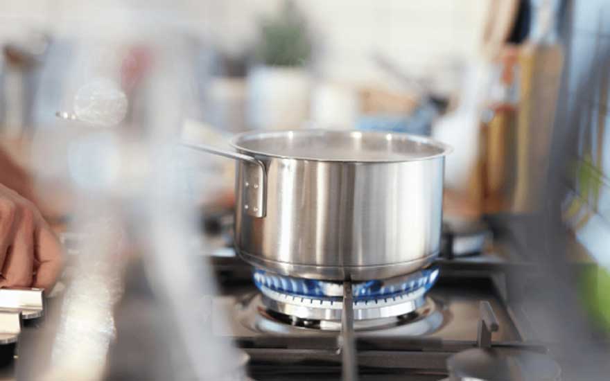 Een roestvrijstalen pan kookt op een gaspit met blauwe vlammen, terwijl een hand de knop van het fornuis voorzichtig bedient.
