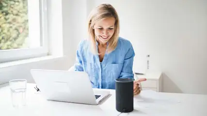Vrouw achter laptop met glas water en zwarte slimme speaker op wit bureau bij een raam.