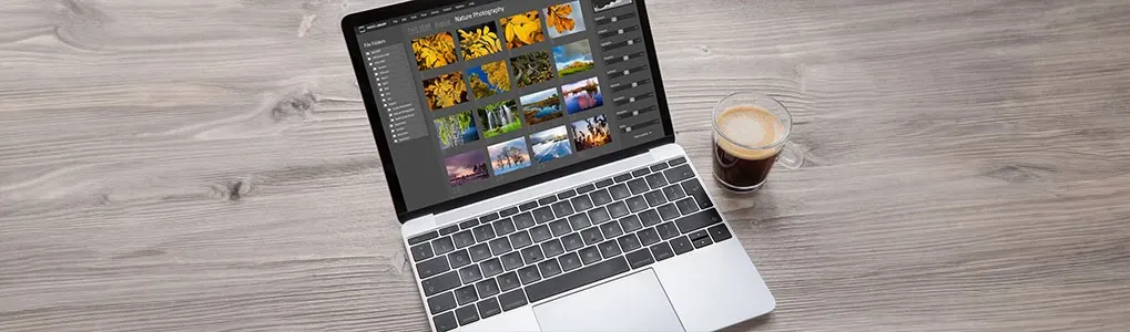 Laptop displays photo editing software next to a glass of coffee on a wood-look surface.