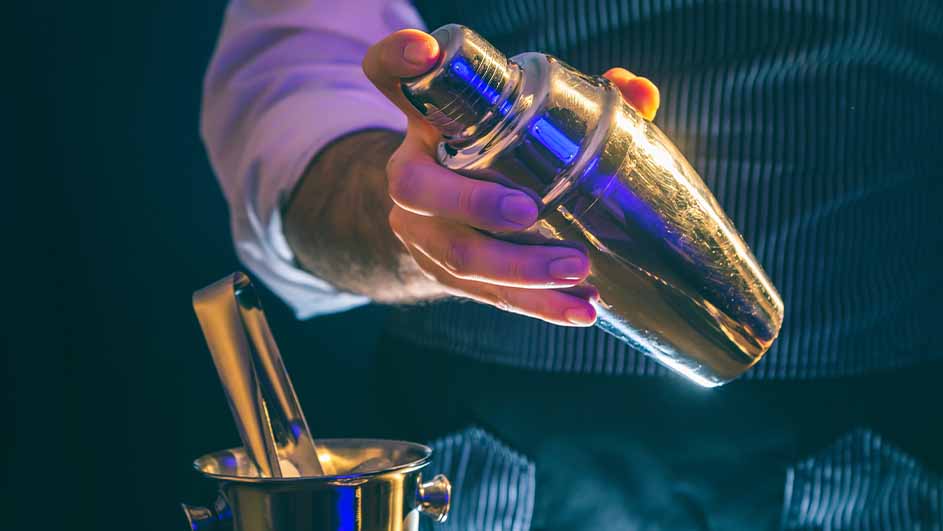 Een hand schudt een zilveren cocktailshaker. IJstang in een zilveren emmer is te zien. De achtergrond is donkerblauw.