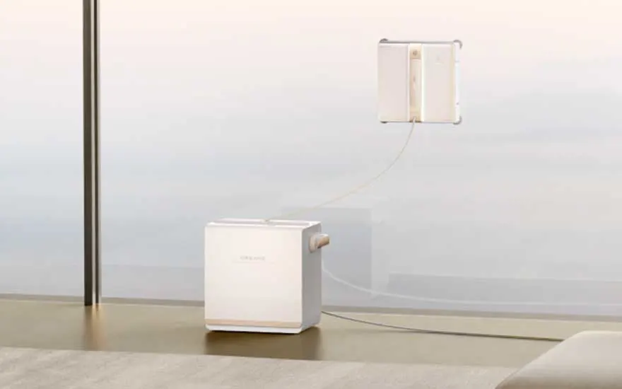Een wit compact apparaat staat op de vloer, verbonden met een wandmodule via dunne kabels in een minimalistische ruimte.